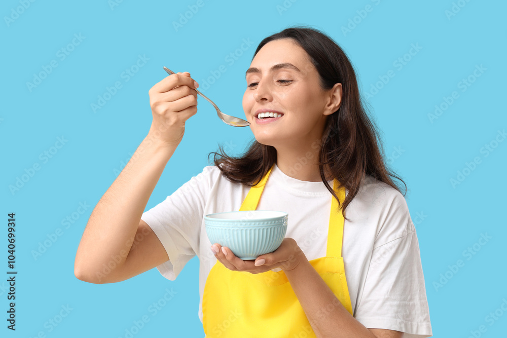 Young woman in apron eating tasty soup on blue background Young woman in apron eating tasty soup on blue background