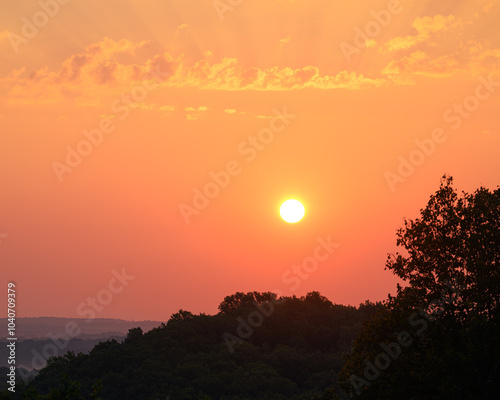 A brilliant orange sun rising over the forested hills of southern indiana. Beautiful and serene scene. Simple composition with sun, clouds, and forest photo