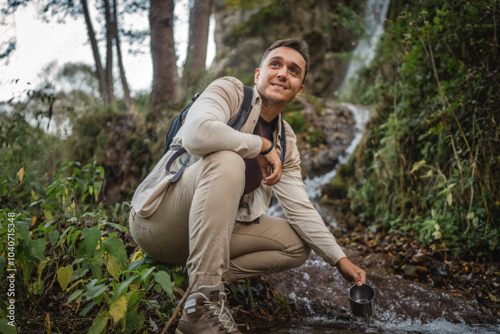 Adult man hiker backpacker squats by a river, filling a mug with water Adult man hiker backpacker squats by a river, filling a mug with water