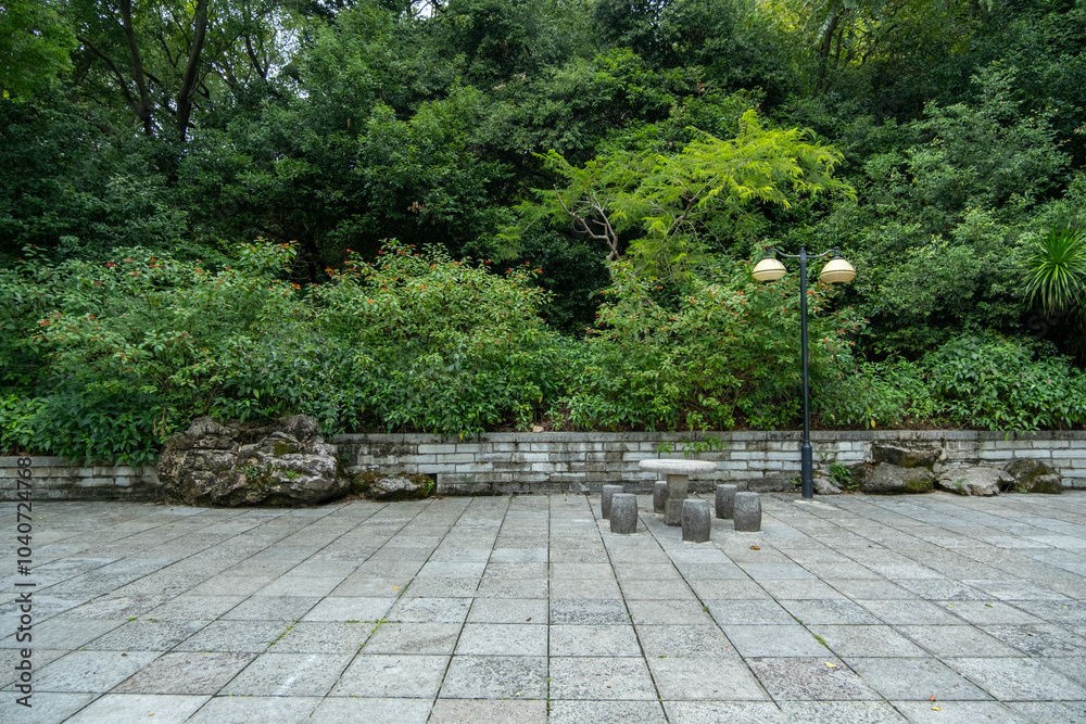 Background texture of an outdoor space with a stone paving surrounded by lush greenery.A low retaining wall separates the public ground from the plants and trees.a hardscape in a Chinese or Asian park Background texture of an outdoor space with a stone paving surrounded by lush greenery.A low retaining wall separates the public ground from the plants and trees.a hardscape in a Chinese or Asian park