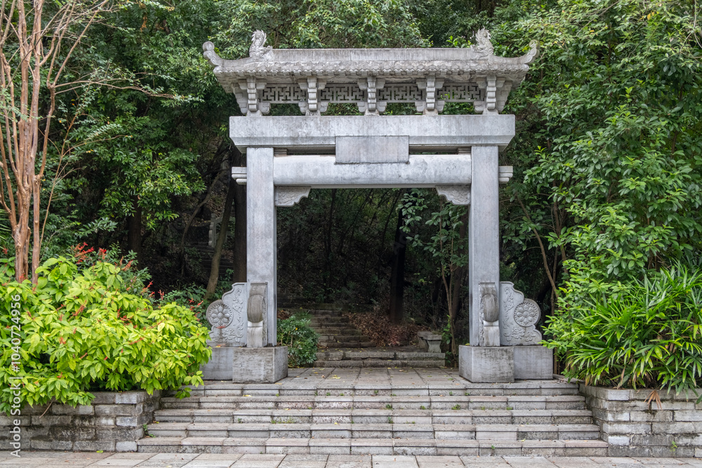 Empty space on stone plaque. A traditional Chinese stone archway with a curved roof and intricate carvings leads to a pathway to a forested hill with steps that lead up to a hiking trail in a park Empty space on stone plaque. A traditional Chinese stone archway with a curved roof and intricate carvings leads to a pathway to a forested hill with steps that lead up to a hiking trail in a park