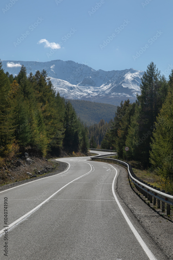 Long curved asphalt road running along the mountain pass. Crash barrier are along the road. Golden autumn roadscape with yellow trees and wild nature. Snowy peaks in the distance Long curved asphalt road running along the mountain pass. Crash barrier are along the road. Golden autumn roadscape with yellow trees and wild nature. Snowy peaks in the distance