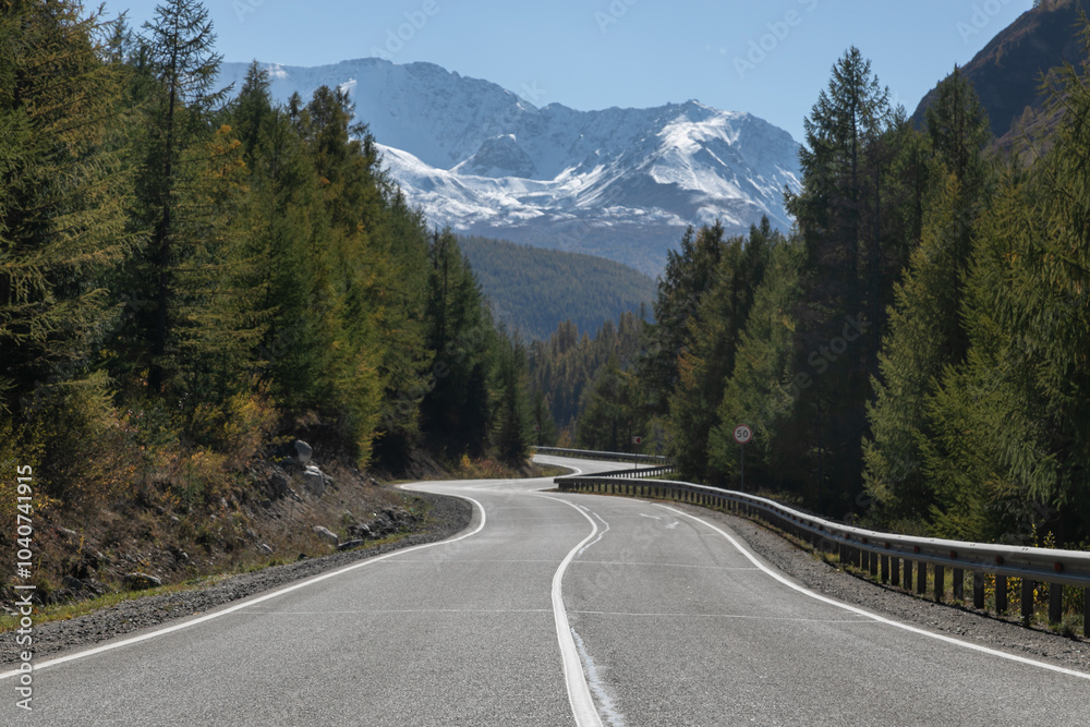 Long curved asphalt road running along the mountain pass. Crash barrier are along the road. Golden autumn roadscape with yellow trees and wild nature. Snowy peaks in the distance Long curved asphalt road running along the mountain pass. Crash barrier are along the road. Golden autumn roadscape with yellow trees and wild nature. Snowy peaks in the distance