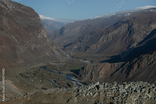 Panoramic landscape of a mountain pass from above. Long curved road leading from the peak to the wide valley. Mountain river streams through the valley. Golden autumn nature: September in Altai.