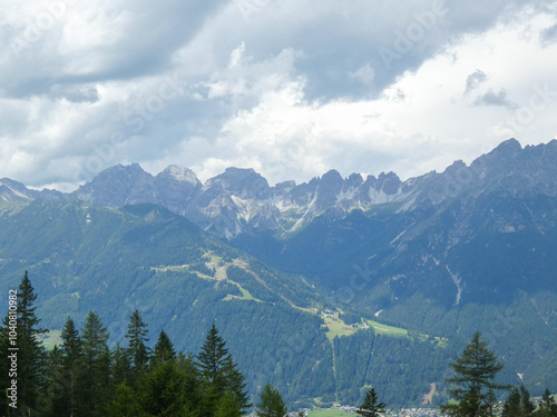 The Stubai Valley showcases the striking peaks of the Austrian Alps beneath a dramatic sky. Tall pine trees enhance the view, highlighting the natural landscape of the Tyrol region