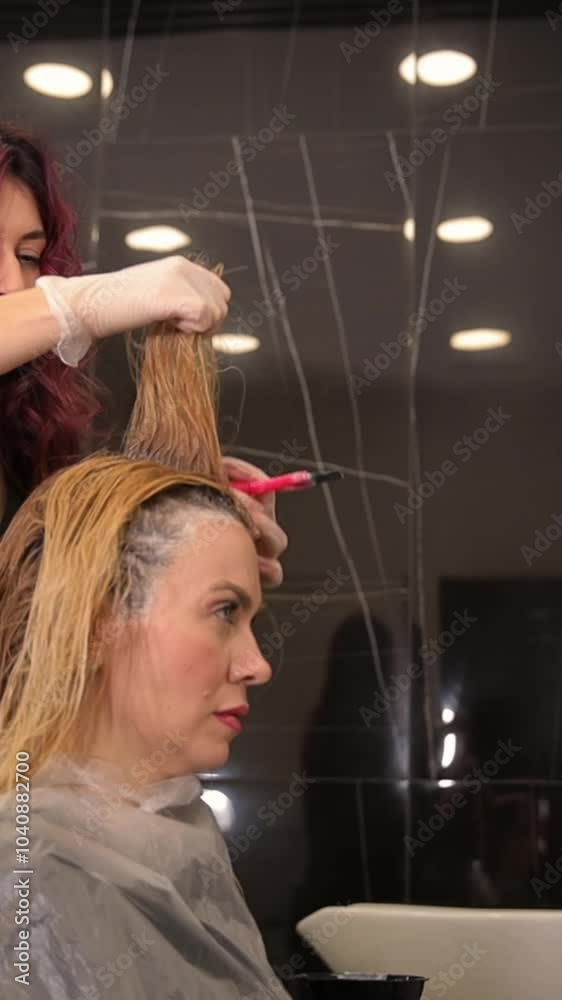A hairdresser paints the roots of blond hair with a brush for a middle-aged woman. Vertical video A hairdresser paints the roots of blond hair with a brush for a middle-aged woman. Vertical video