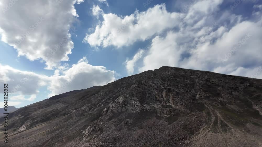 Cinematic Side View of a Majestic Mountain Range with Dark Peaks, Rocky Outcrops,Grass Patches.Below,Winding Black Bitumen Road Leads into a Deep Valley.Above,Blue Sky with Fluffy Clouds,Rain Falls Cinematic Side View of a Majestic Mountain Range with Dark Peaks, Rocky Outcrops,Grass Patches.Below,Winding Black Bitumen Road Leads into a Deep Valley.Above,Blue Sky with Fluffy Clouds,Rain Falls