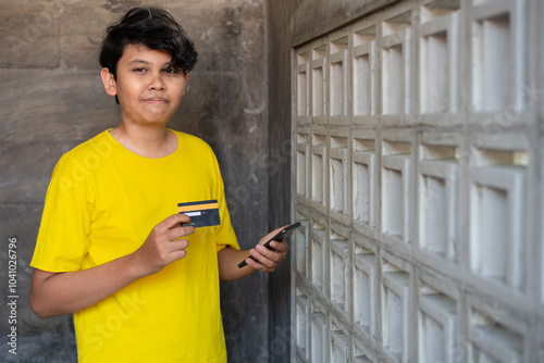 A young Indonesian man wearing a yellow shirt is holding a credit card and his smartphone and is about to make a purchase transaction, business and finance concepts photo