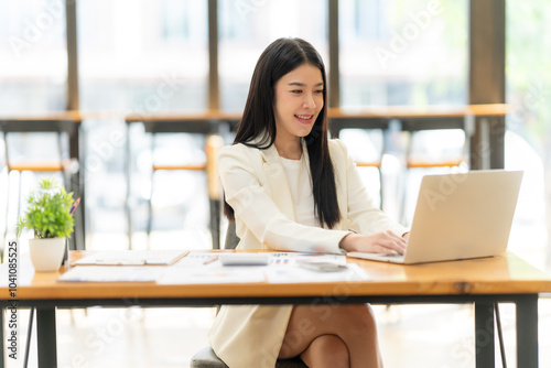 Focused and Driven: A young Asian businesswoman in a stylish white blazer, engrossed in her work on a laptop. Her focused expression and confident posture suggest dedication and ambition.