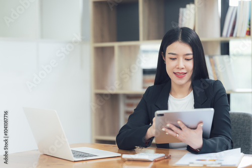 Confident Businesswoman Using Tablet: A professional young woman in a business suit smiles as she works on a tablet in a modern office setting, showcasing focus, technology.