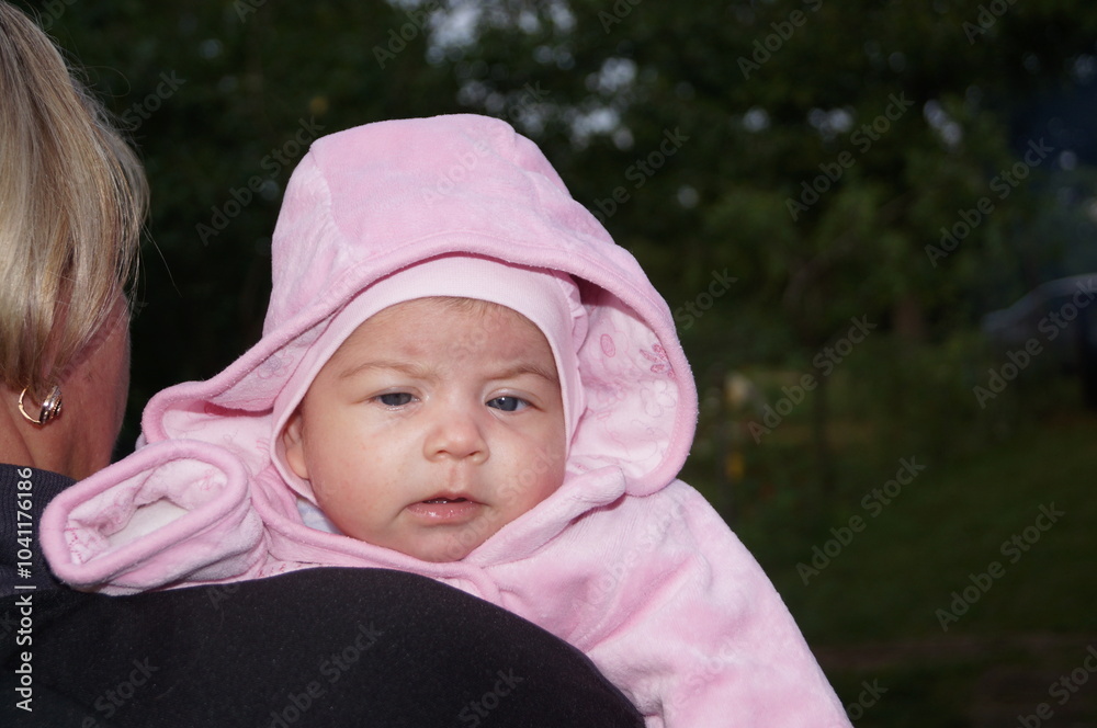 A woman holds a newborn, baby, toddler or small child with a hood over his head, who is looking towards the camera and squinting. A woman holds a newborn, baby, toddler or small child with a hood over his head, who is looking towards the camera and squinting.
