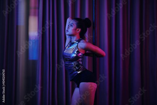 Asian woman in metallic dance outfit standing by pole in dimly lit room, reflecting on her practice. Curtains create backdrop for serene and focused moment