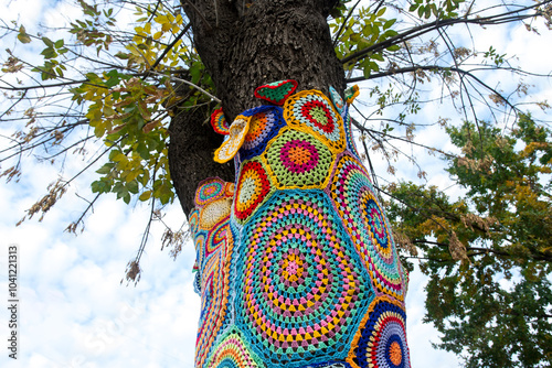A tree covered in colorful crocheted patterns as part of urban yarn bombing, with vibrant geometric shapes and textures. The creative installation stands out against the natural and urban backdrop