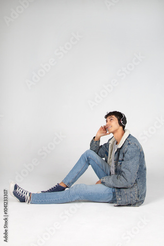 Chico joven vestido de jeans con auriculares, escuchando música o podcast, sentado en el suelo mirando al cielo. Publicidad, cartelería, ofertas. photo