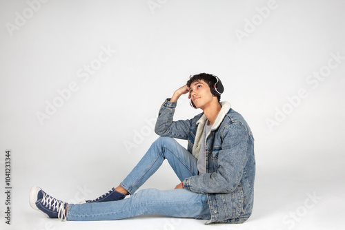 Chico joven vestido de jeans con auriculares, escuchando música o podcast, sentado en el suelo mirando al cielo. Publicidad, cartelería, ofertas.