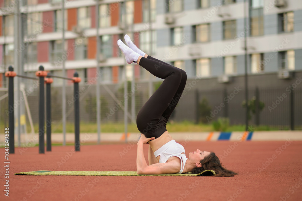 Fototapeta premium fit woman lifts her legs into the air while practicing a shoulder stand on a yoga mat in an urban park. The photo highlights the importance of core strength and balance in contemporary outdoor