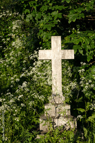 White stone cross memorial in old churchyard situated in bushes with wild flowers.