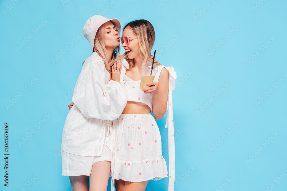 Two young beautiful smiling blond hipster female in trendy summer clothes. Carefree women posing near blue wall in studio. Positive models having fun. Cheerful and happy Two young beautiful smiling blond hipster female in trendy summer clothes. Carefree women posing near blue wall in studio. Positive models having fun. Cheerful and happy