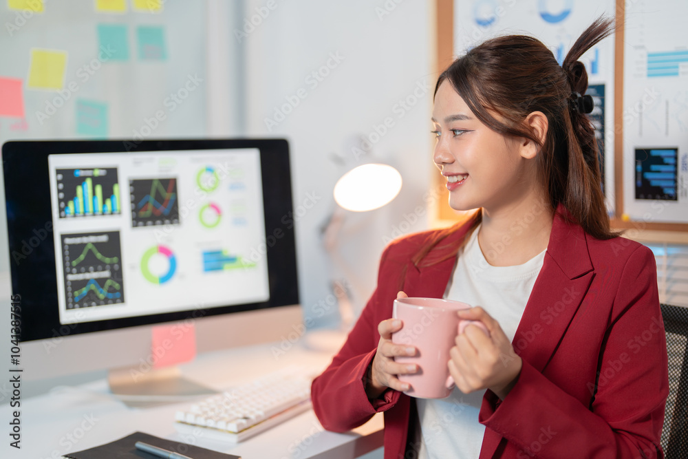 Young asian businesswoman is holding a mug in her hands and smiling at something off-camera while sitting at her desk in the office Young asian businesswoman is holding a mug in her hands and smiling at something off-camera while sitting at her desk in the office