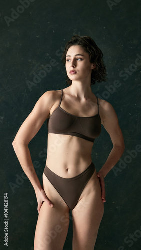 Young woman stands gracefully in neutral-toned bikini, gazing off to side against dark green studio background. Beauty and calm. Concept of spa procedure, self-care treatments, self-love, style. Ad