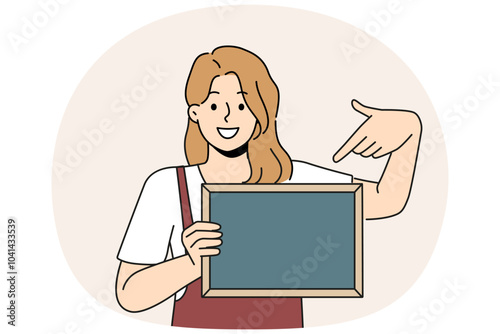 Woman waiter is holding chalkboard with copy space for drawing menu or promotional offers from restaurant. Cheerful girl waiter in apron points with finger at place for name of bar or cafe logo