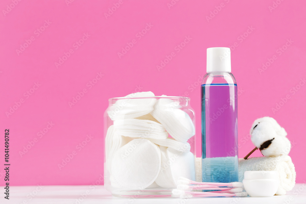 Cotton pads in glass jar with flower and bottle of liquid on pink background