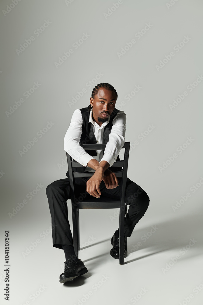 A young African American man in a stylish suit exudes confidence while seated on a chair against a grey background. A young African American man in a stylish suit exudes confidence while seated on a chair against a grey background.