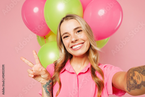 Close up young woman in shirt celebrating birthday holiday near bunch of colorful air balloons do selfie shot pov on mobile cell phone show v-sign isolated on plain pink background Lifestyle concept