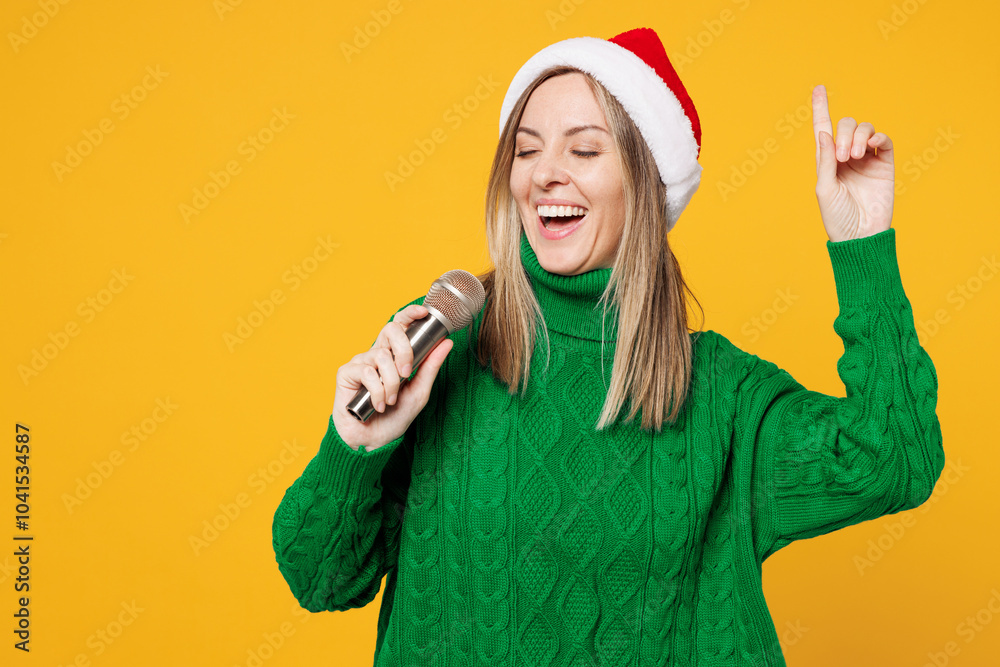 Merry fun young woman wear green warm cozy sweater Santa hat posing sing song in microphone point finger up isolated on plain yellow background. Happy New Year, celebration Christmas holiday concept. Merry fun young woman wear green warm cozy sweater Santa hat posing sing song in microphone point finger up isolated on plain yellow background. Happy New Year, celebration Christmas holiday concept.