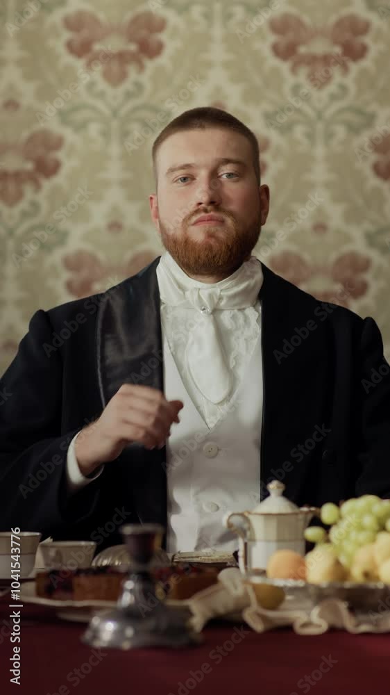Vertical waist up portrait of young bearded aristocrat looking at camera while adjusting white tie, sitting at table in classically decorated dining room at daytime Vertical waist up portrait of young bearded aristocrat looking at camera while adjusting white tie, sitting at table in classically decorated dining room at daytime