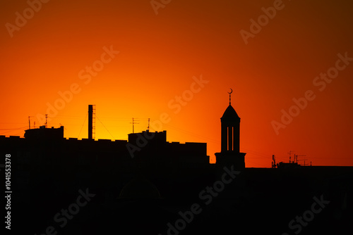 Silhouettes of buildings and a mosque against an orange-red sunset with a clear sky