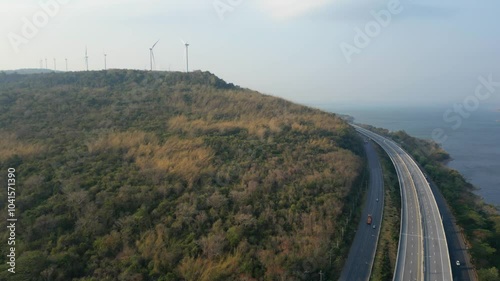 Aerial view of a highway winding through a hilly, forested landscape with wind turbines in the distance, near a coastline.

