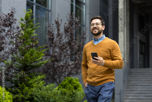 Young smiling businessman walking outside of office building, hand in pocket, holding mobile phone and looking forward with satisfaction