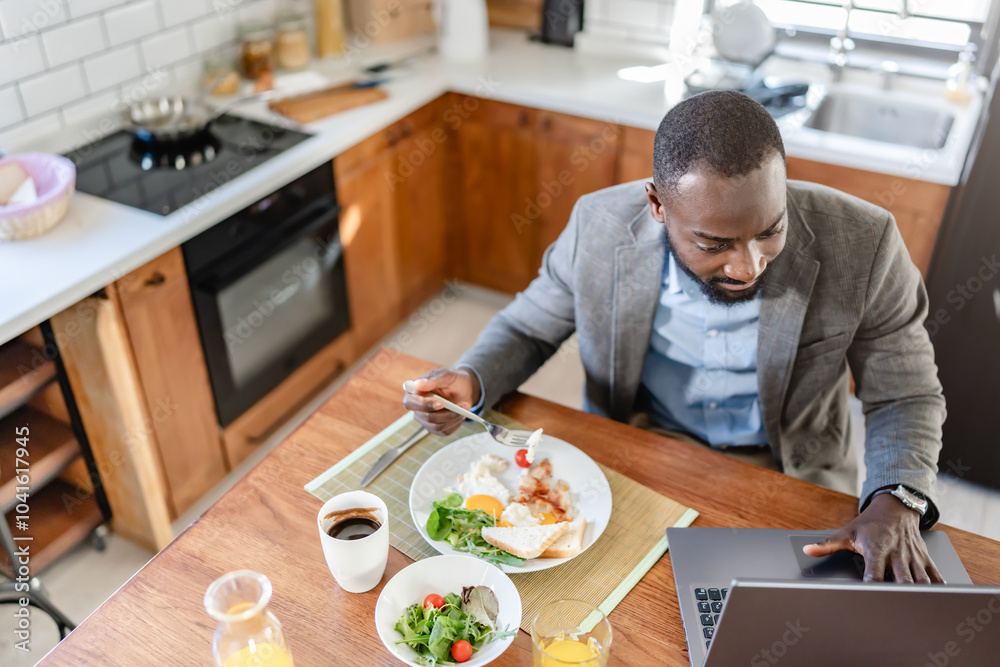 African-American man working on a laptop while eating breakfast at home. He is multitasking in a bright, modern kitchen, combining work and a healthy meal. African-American man working on a laptop while eating breakfast at home. He is multitasking in a bright, modern kitchen, combining work and a healthy meal.