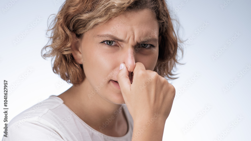 A young woman with short curly hair and a white t-shirt pinches her nose in clear discomfort, reacting to a bad smell. Her facial expression vividly conveys the unpleasantness of the odor. Ideal for A young woman with short curly hair and a white t-shirt pinches her nose in clear discomfort, reacting to a bad smell. Her facial expression vividly conveys the unpleasantness of the odor. Ideal for