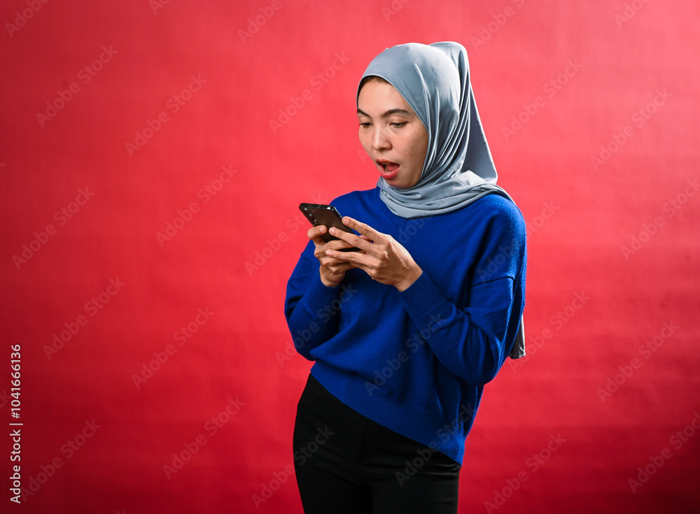 Asian woman in a grey hijab and blue sweater holds a smartphone, looking surprised as she covers her mouth with one hand. She stands against a bright red background, reacting to something Asian woman in a grey hijab and blue sweater holds a smartphone, looking surprised as she covers her mouth with one hand. She stands against a bright red background, reacting to something