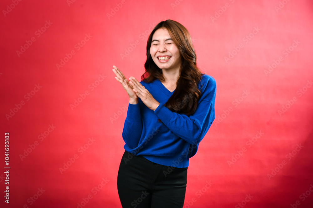 An Asian woman in a blue sweater, clapping her hands with a cheerful expression, standing against a red background. Her joyful energy and excitement are vividly displayed. An Asian woman in a blue sweater, clapping her hands with a cheerful expression, standing against a red background. Her joyful energy and excitement are vividly displayed.