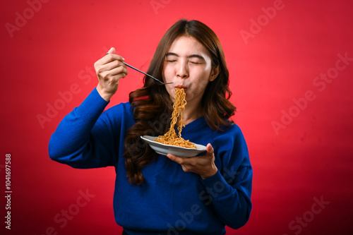 An Asian woman in a blue sweater happily slurps noodles from a plate, her eyes closed in enjoyment. She holds a fork and stands against a vibrant red background, savoring the delicious meal. photo