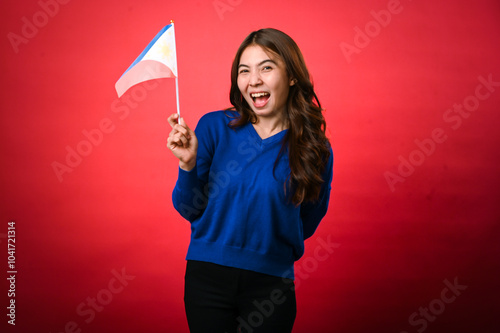 An Asian woman holding a small Philippine flag in her right hand, smiling joyfully. She wears a blue sweater against a red background. Her long hair cascades over her shoulders