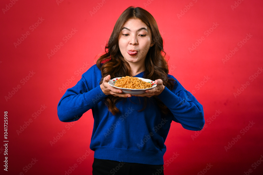 An Asian woman in a blue sweater holds a plate of noodles close to her face, licking her lips in anticipation. She stands against a red background with a playful and hungry expression An Asian woman in a blue sweater holds a plate of noodles close to her face, licking her lips in anticipation. She stands against a red background with a playful and hungry expression