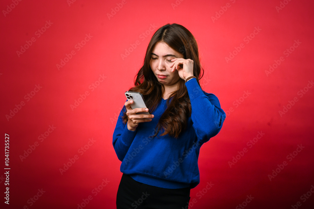 An Asian woman in a blue sweater frowns while looking at her phone, appearing confused or displeased. She stands against a bright red background, holding the phone with both hands An Asian woman in a blue sweater frowns while looking at her phone, appearing confused or displeased. She stands against a bright red background, holding the phone with both hands