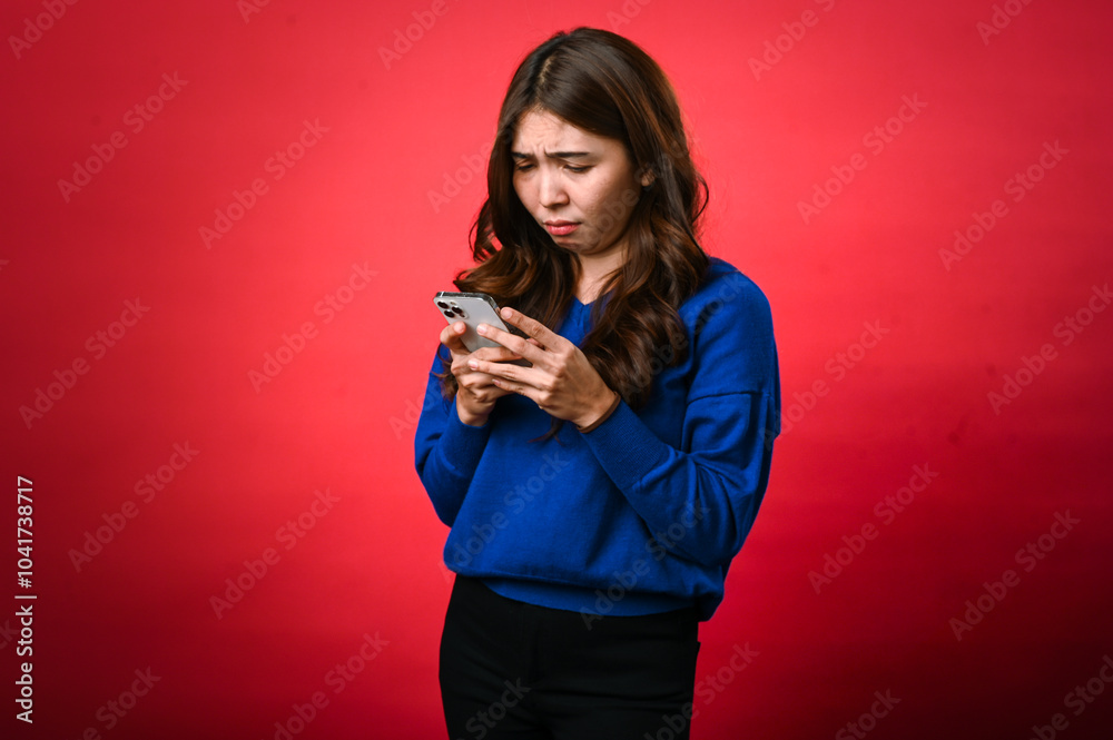 An Asian woman in a blue sweater frowns while looking at her phone, appearing confused or displeased. She stands against a bright red background, holding the phone with both hands An Asian woman in a blue sweater frowns while looking at her phone, appearing confused or displeased. She stands against a bright red background, holding the phone with both hands