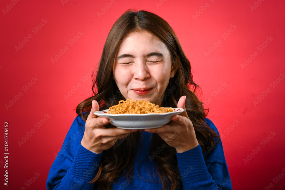 An Asian woman in a blue sweater holds a plate of noodles close to her face, licking her lips in anticipation. She stands against a red background with a playful and hungry expression An Asian woman in a blue sweater holds a plate of noodles close to her face, licking her lips in anticipation. She stands against a red background with a playful and hungry expression