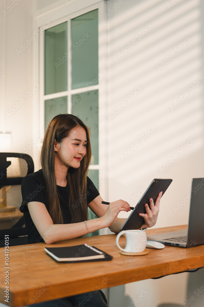 young woman is working on tablet at wooden desk, smiling as she engages with her device. laptop and notebook are also present, creating productive workspace young woman is working on tablet at wooden desk, smiling as she engages with her device. laptop and notebook are also present, creating productive workspace