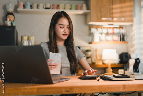 young woman is focused on her work at wooden desk, holding receipt while using laptop and calculator. cozy workspace is well organized, reflecting productive atmosphere