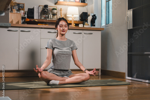 woman practices meditation in cozy indoor space, sitting cross legged on yoga mat. She exudes calmness and tranquility, surrounded by minimalist kitchen setting