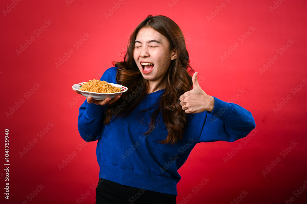 An Asian woman wearing a blue sweater smiles enthusiastically while holding a plate of noodles in one hand and giving a thumbs-up with the other. She stands against a bright red background An Asian woman wearing a blue sweater smiles enthusiastically while holding a plate of noodles in one hand and giving a thumbs-up with the other. She stands against a bright red background