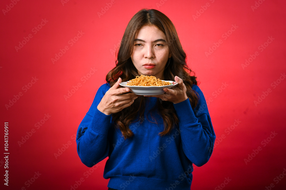 An Asian woman in a blue sweater holds a plate of noodles close to her face, licking her lips in anticipation. She stands against a red background with a playful and hungry expression An Asian woman in a blue sweater holds a plate of noodles close to her face, licking her lips in anticipation. She stands against a red background with a playful and hungry expression