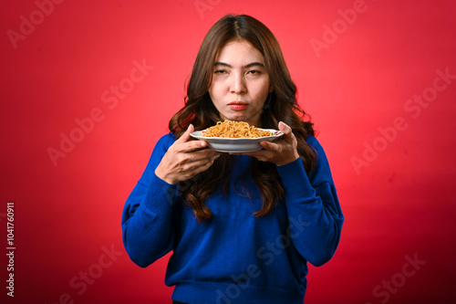 An Asian woman in a blue sweater holds a plate of noodles close to her face, licking her lips in anticipation. She stands against a red background with a playful and hungry expression photo