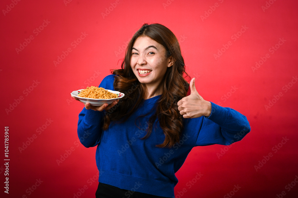 An Asian woman wearing a blue sweater smiles enthusiastically while holding a plate of noodles in one hand and giving a thumbs-up with the other. She stands against a bright red background An Asian woman wearing a blue sweater smiles enthusiastically while holding a plate of noodles in one hand and giving a thumbs-up with the other. She stands against a bright red background
